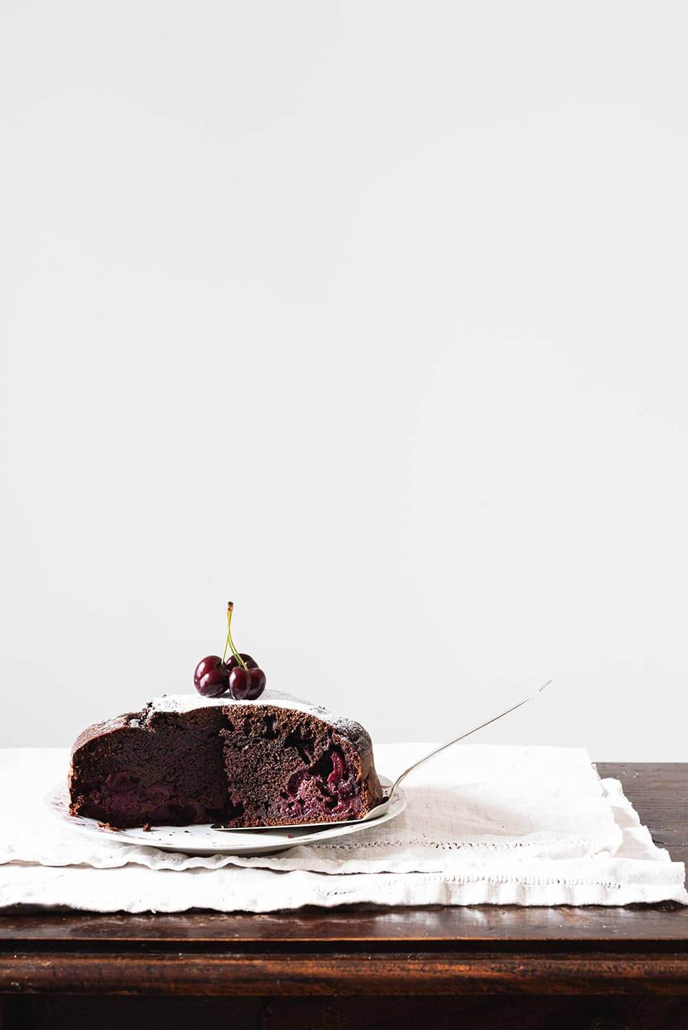 Bizcocho de chocolate con cerezas esponjoso En una mesa cubierta con un mantel blanco. encima está un plato blanco. En el plato está el bizcocho de cerezas y chocolate. Ya está cortada. Hay una pala de tartas que también es el plato.