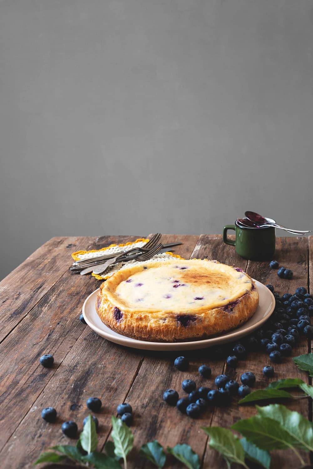 Cheesecake de arándanos con horno En una mesa de madera hay un plato blanco. En el plato está la tarta de queso de arándanos. Todavía no está cubierta con la salsa de arándanos. Alrededor hay hojas, arándanos, una taza con la salsa y cubiertos.