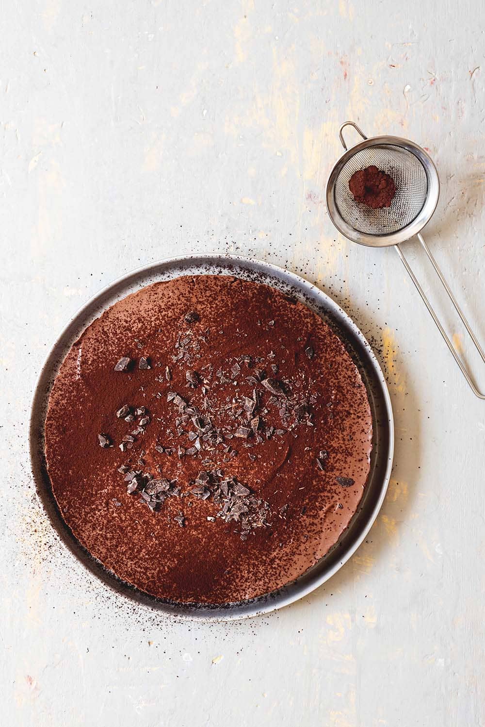 Cheesecake de chocolate sin horno En una mesa gris está un plato gris. Encima está el Cheesecake de chocolate. Al lado hay un colador con cacao en polvo.