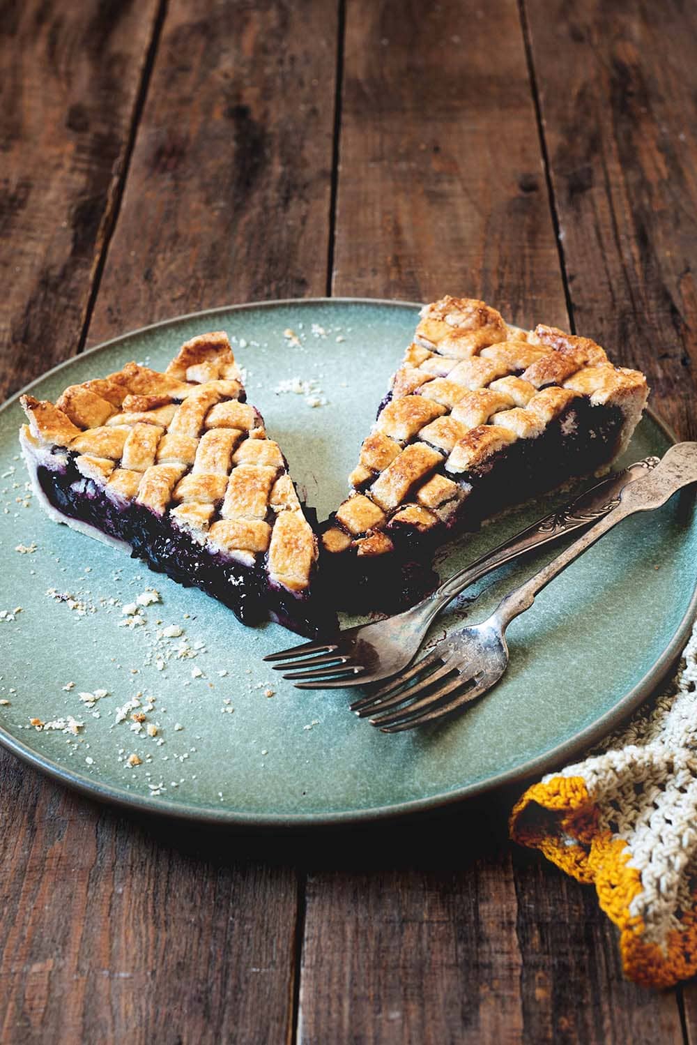 Cherry pie En una mesa de madera está puesta un plato azul. Encima están dos raciones de Cherry pie. Al lado hay dos tenedores y un paño de cocina.