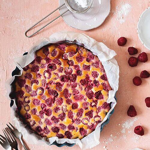 En una mesa de color rosa clara está en un molde el clafoutis de frambuesas. Alrededor hay cubiertos, frambuesas frescas y un colador con azúcar glas.