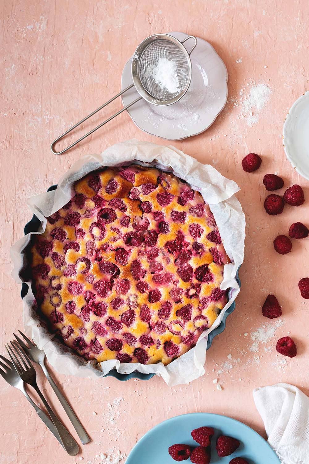 Clafoutis de frambuesas fácil y rápido En una mesa de color rosa clara está en un molde el clafoutis de frambuesas. Alrededor hay cubiertos, frambuesas frescas y un colador con azúcar glas.
