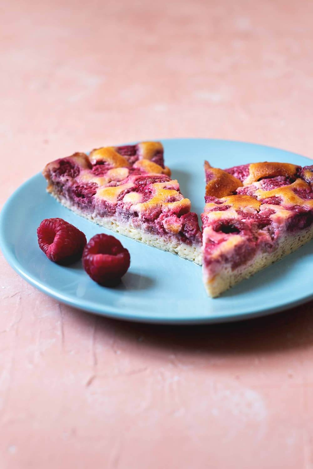 Clafoutis de frambuesas En una mesa de color rosa clara hay un plato azul. En el plato hay dos raciones de clafoutis de frambuesas. Alrededor hay dos frambuesas frescas.