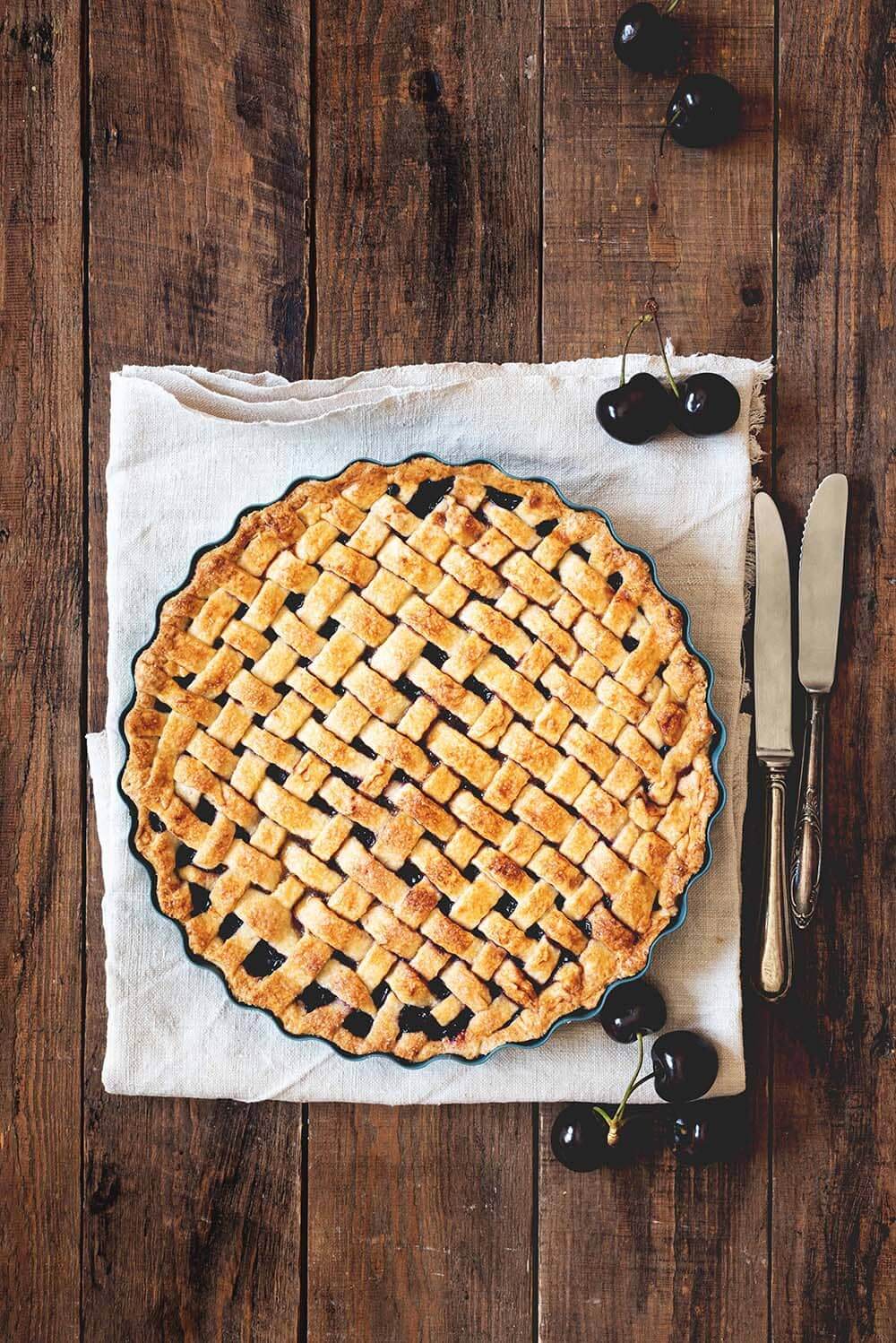 Clásica tarta americana de cerezas En una mesa de madera está puesta la clásica tarta americana de cerezas. Abajo de la tarta hay un paño de cocina blanco. Alrededor de la tarta hay cerezas frescas.