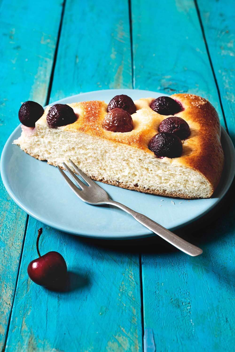 Coca de cerezas En una mesa de color azul está un plato azul. Encima está una ración de coca de cerezas. Se ve muy esponjoso.