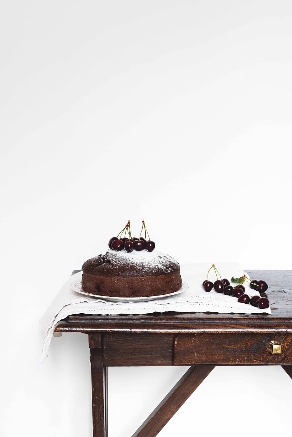 Cómo hacer bizcocho de chocolate con cerezas esponjoso En una mesa cubierta con un mantel blanco. encima está un plato blanco. En el plato está el bizcocho de cerezas y chocolate . Todavía está sin cortar. Al lado hay un puñado de cerezas.