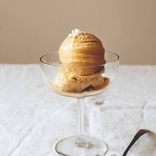 Una mesa está cubierta con un mantel blanco. En una copa de cristal hay dos bolas de helado caramelo casero.