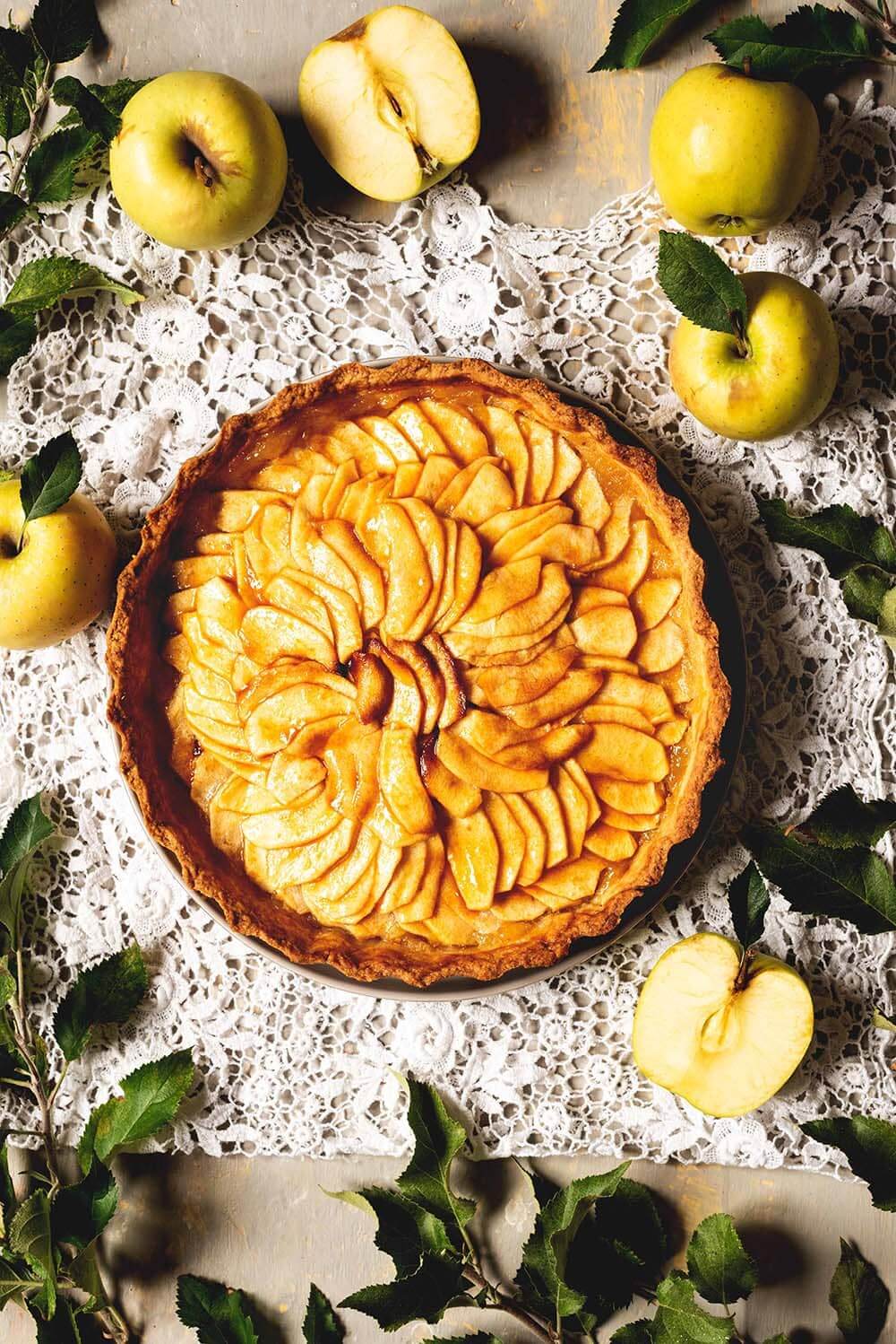 Una mesa gris está cubierta con un mantel blanco vintage. Encima está la tarta de manzana clásica francesa. Alrededor hay manzanas y hojas verdes de un manzano.