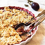 Una mesa está cubierta con un mantel blanco. Encima está el molde de cerámica con el crumble de ciruelas. Se ve jugoso. En el molde hay dos cucharas.