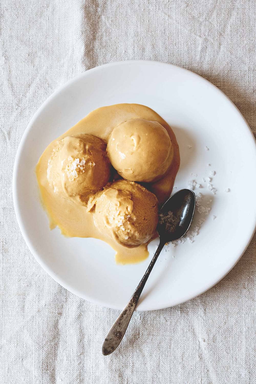 Una mesa está cubierta con un mantel blanco. Encima hay un plato lanco. En el plato hay tres bolas de helado caramelo casero. Al lado hay una cuchara vintage.