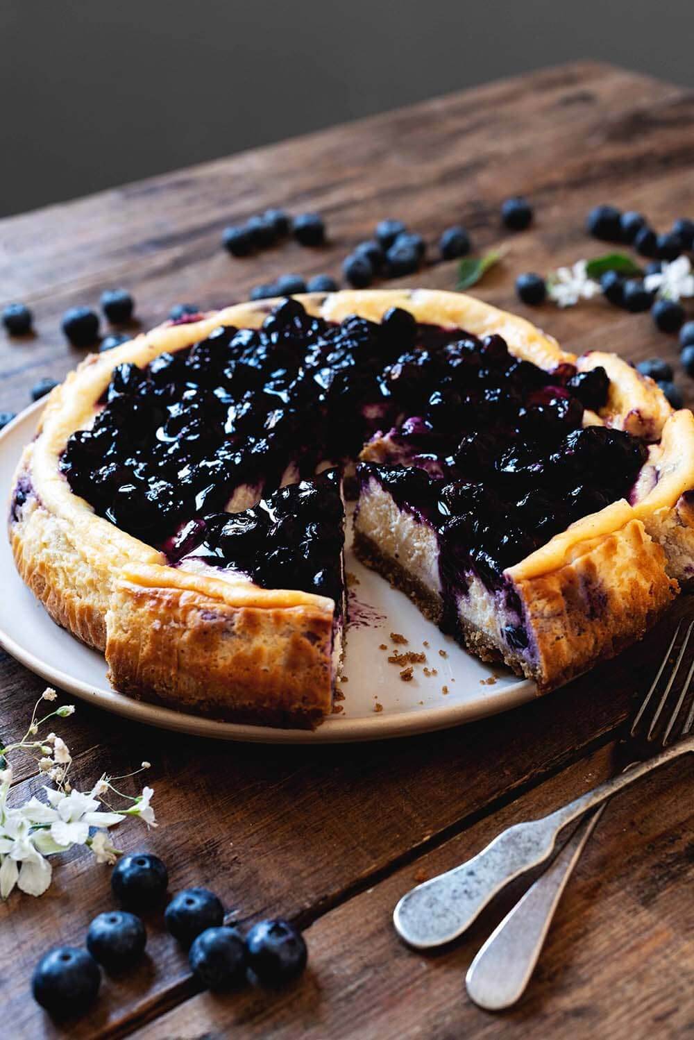 La mejor tarta de queso con arándanos En una mesa de madera hay un plato blanco. En el plato está la tarta de queso de arándanos. Está cortada y está cubierta con la sala de arándanos. Alrededor hay cubiertos de flores blancas, arándanos y hojas. Se puede ver el interior.