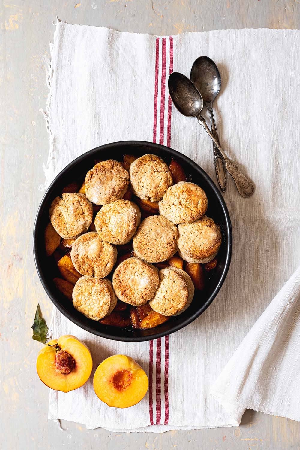 Peach cobbler Una mesa está cubierta con un mantel blanco, Encima hay un molde redondo con el peach cobbler. Alrededor hay un melocotón cortado en la mitad y dos cucharas.