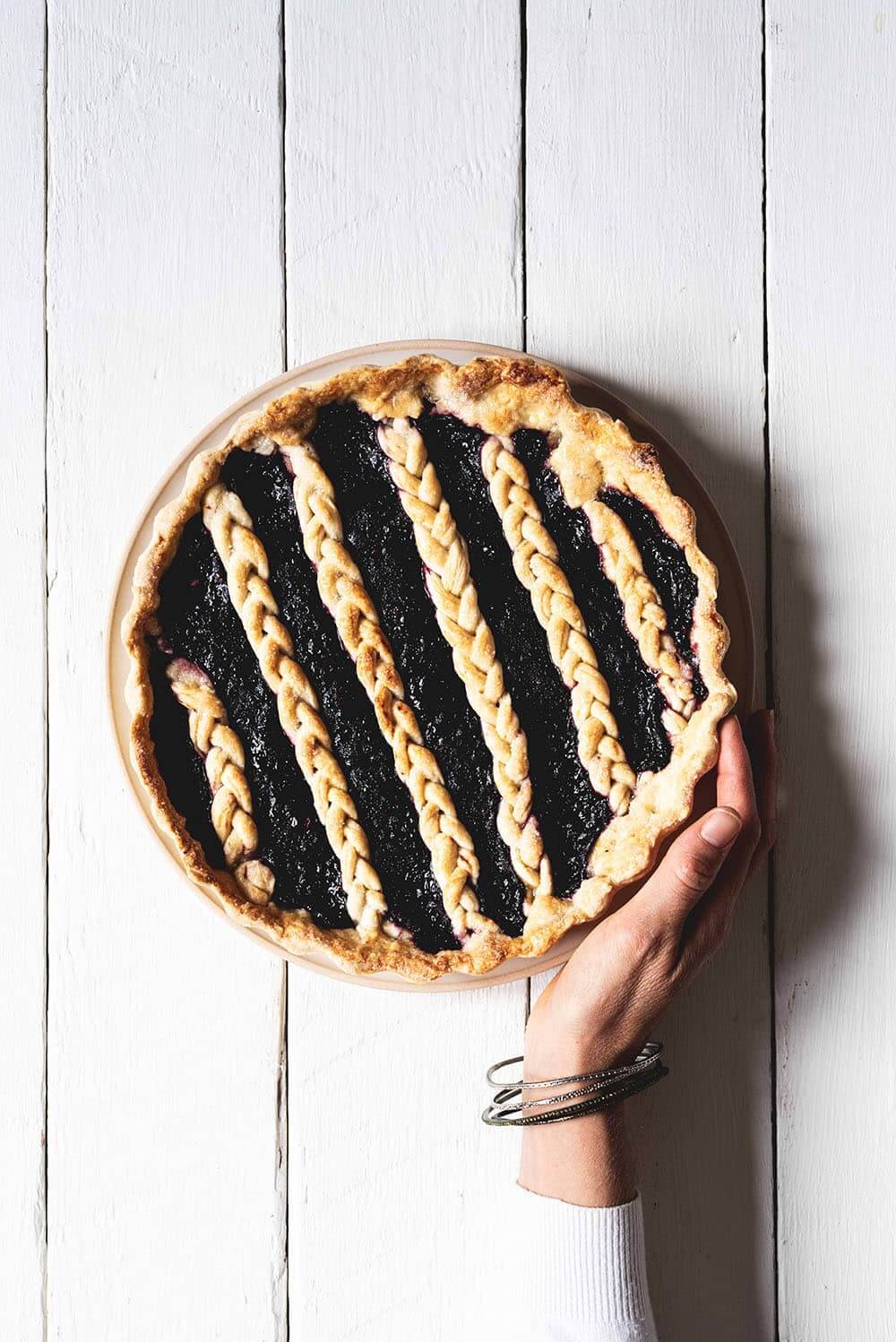 Pie de frutos rojos En una mesa blanca está puesto el Pie de frutos rojos. Una mano está tocando la tarta.