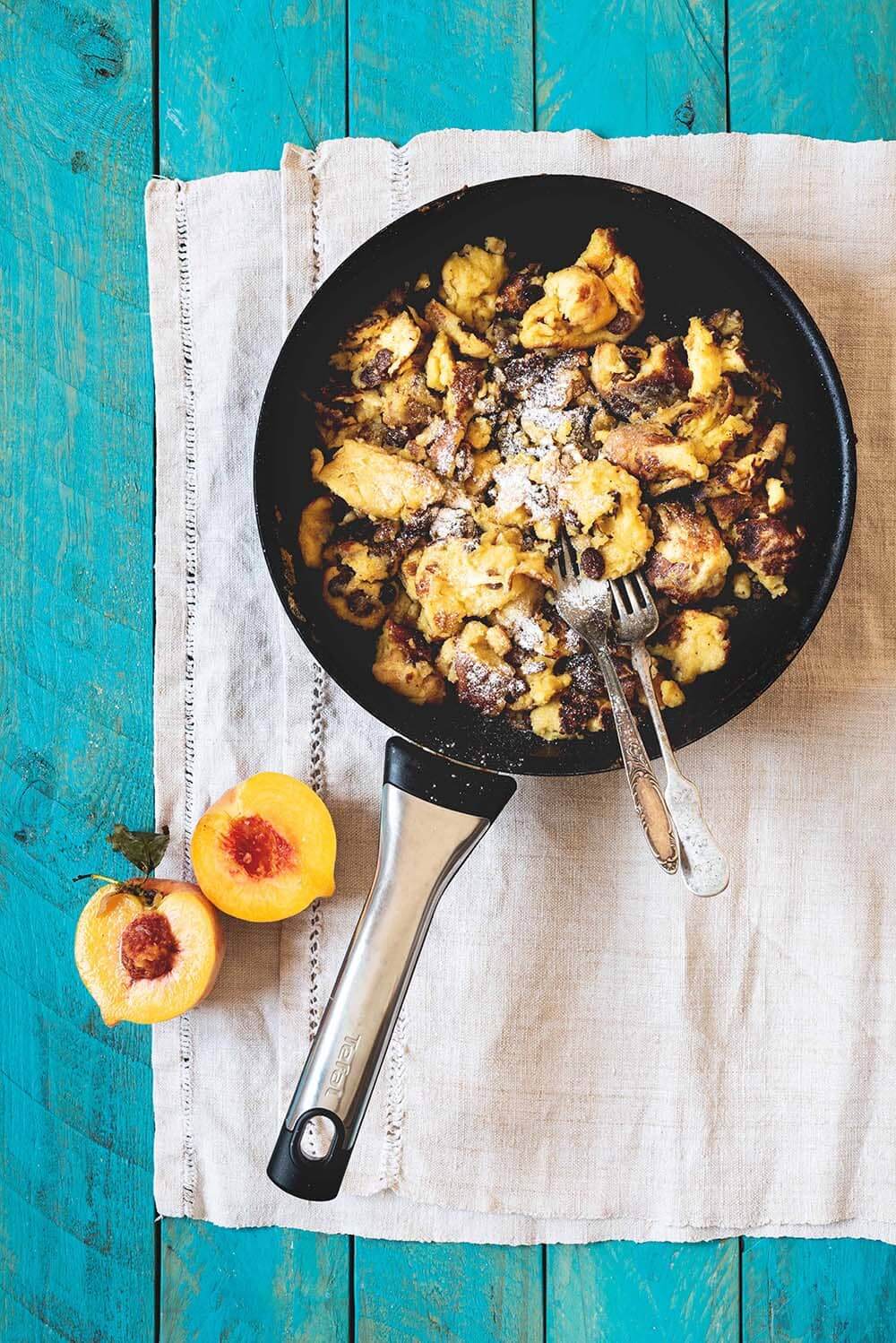 Receta de Kaiserschmarrn Una mesa de madera azul está cubierta con un paño blanco. Encima está la sartén con el Kaiserschmarrn recién hecho. Alrededor hay una fruta fresca.