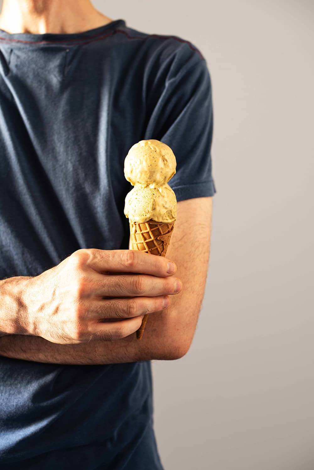 Receta de helado de pistacho cremoso Una persona vestida con una camiseta azul está sujetando en la mano un cucurucho. En el cucurucho hay dos bolas de helado de pistachos cremosos.