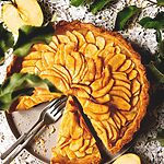 Una mesa gris está cubierta con un mantel blanco vintage. Encima está la tarta de manzana clásica francesa. Ya está cortada. En el molde hay un cuchillo. Alrededor hay manzanas y hojas verdes de un manzano.