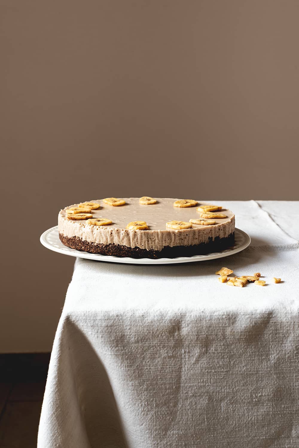 Receta de tarta de queso y plátano sin horno Una mesa gris está cubierta con un mantel blanco vintage. En el borde de la mesa está puesto un plato. Encima está la tarta de queso y plátano sin horno. Alrededor hay migas de chips de plátanos.
