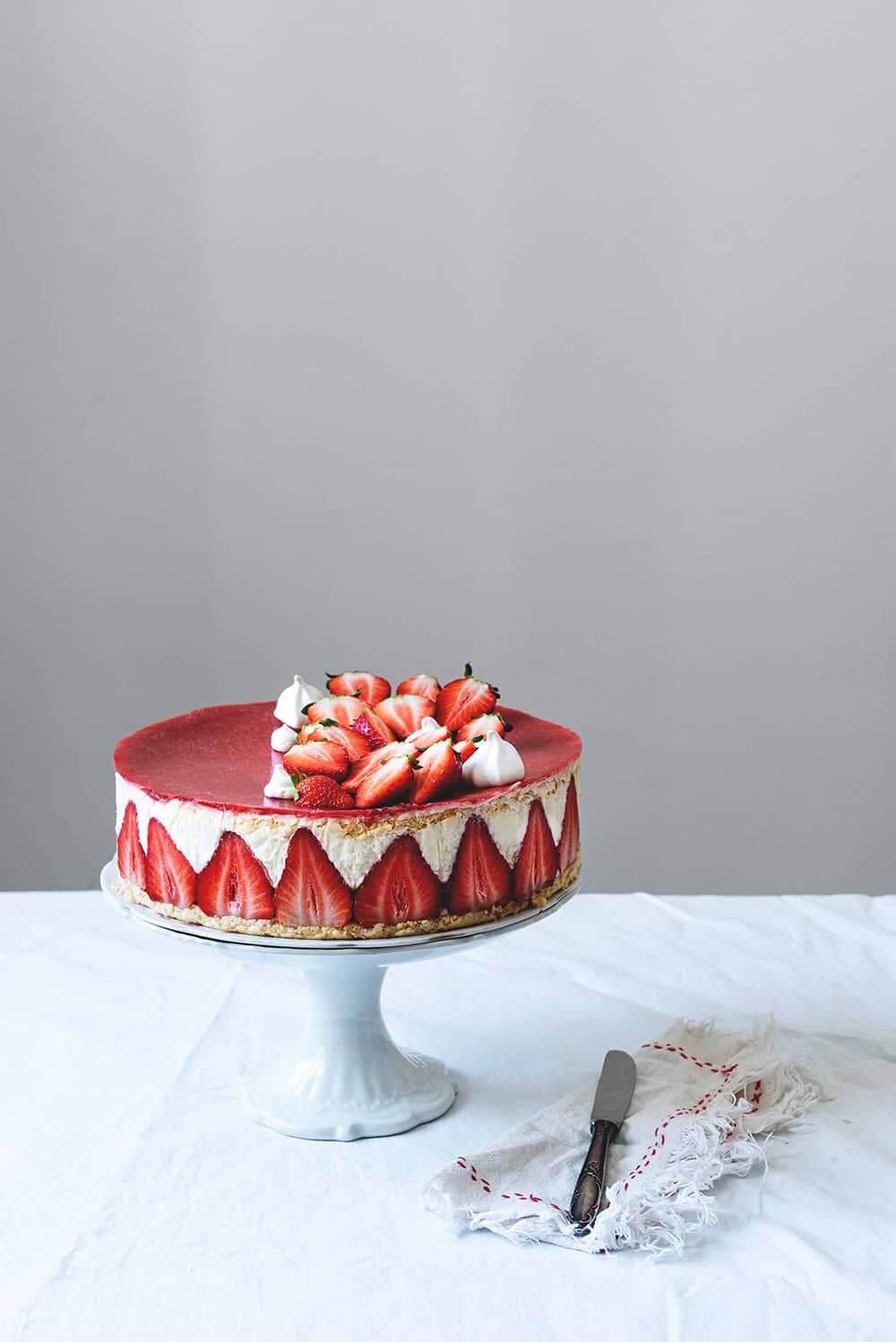 Tarta Fraisier En una mesa cubierta con un mantel blanco. Encima está un portatartas blanco. Encima está la tarta Tarta Fraisier. Al lado hay un cuchillo y una servilleta de algodón.