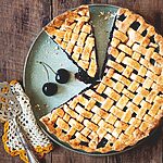 En una mesa de madera está puesta la tarta de cerezas americanas. Alrededor de la tarta hay cerezas frescas, un paño de cocina y una pala de tartas vintage.