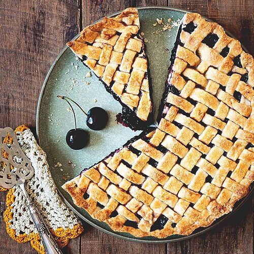 En una mesa de madera está puesta la tarta de cerezas americanas. Alrededor de la tarta hay cerezas frescas, un paño de cocina y una pala de tartas vintage.