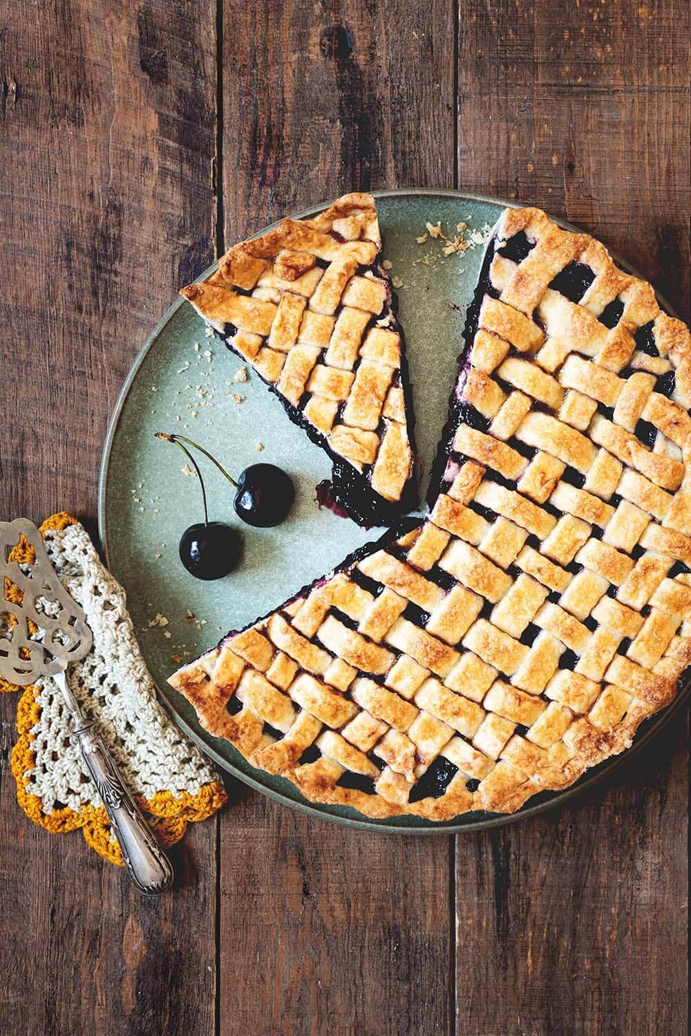 Tarta de cerezas americana En una mesa de madera está puesta la tarta de cerezas americanas. Alrededor de la tarta hay cerezas frescas, un paño de cocina y una pala de tartas vintage.