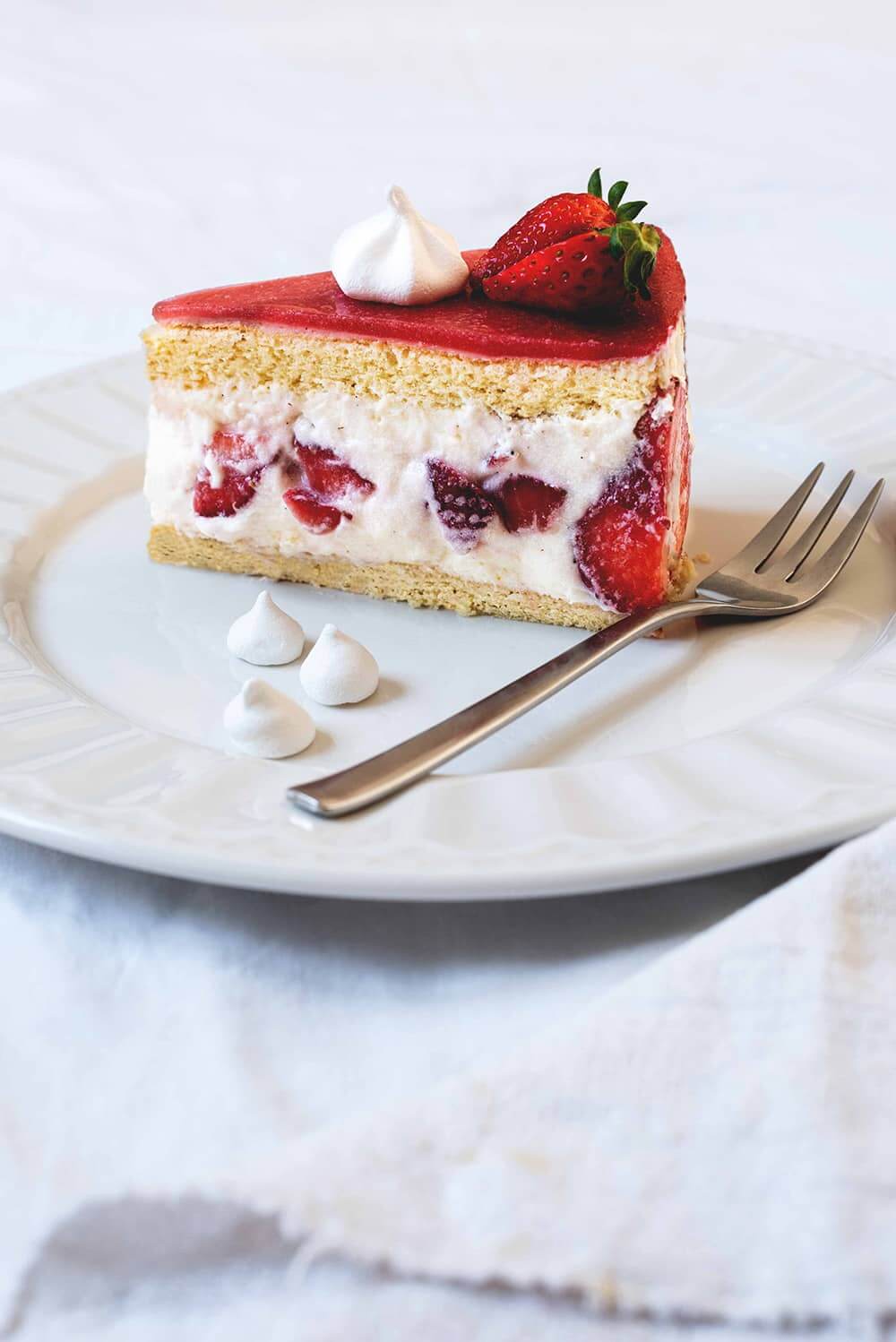 Tarta de fresas francesa En una mesa cubierta con un mantel blanco. Encima está un plato blanco. Encima hay una ración de Tarta Fraisier. Al lado hay un tenedor y un merengue seco.