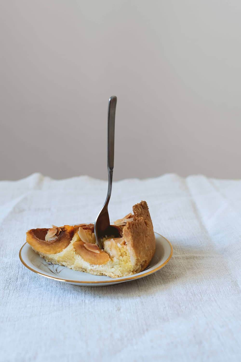 Tarta de nísperos con frangipane En una mesa cubierta con un mantel blanco hay un plato blanco. Encima está una ración de tarta de nísperos. Hay un tenedor pinchado en la tarta.