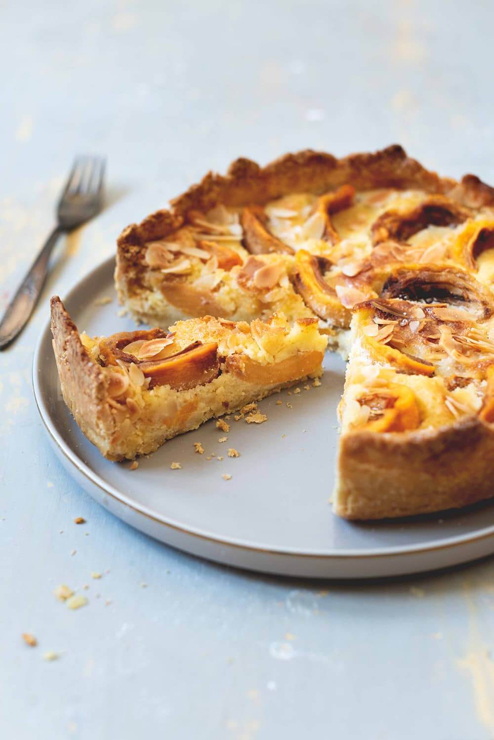 Tarta de nísperos fácil y rico En una mesa de color gris está un plato gris. Encima está la tarta de níspero. Al lado hay un tenedor.