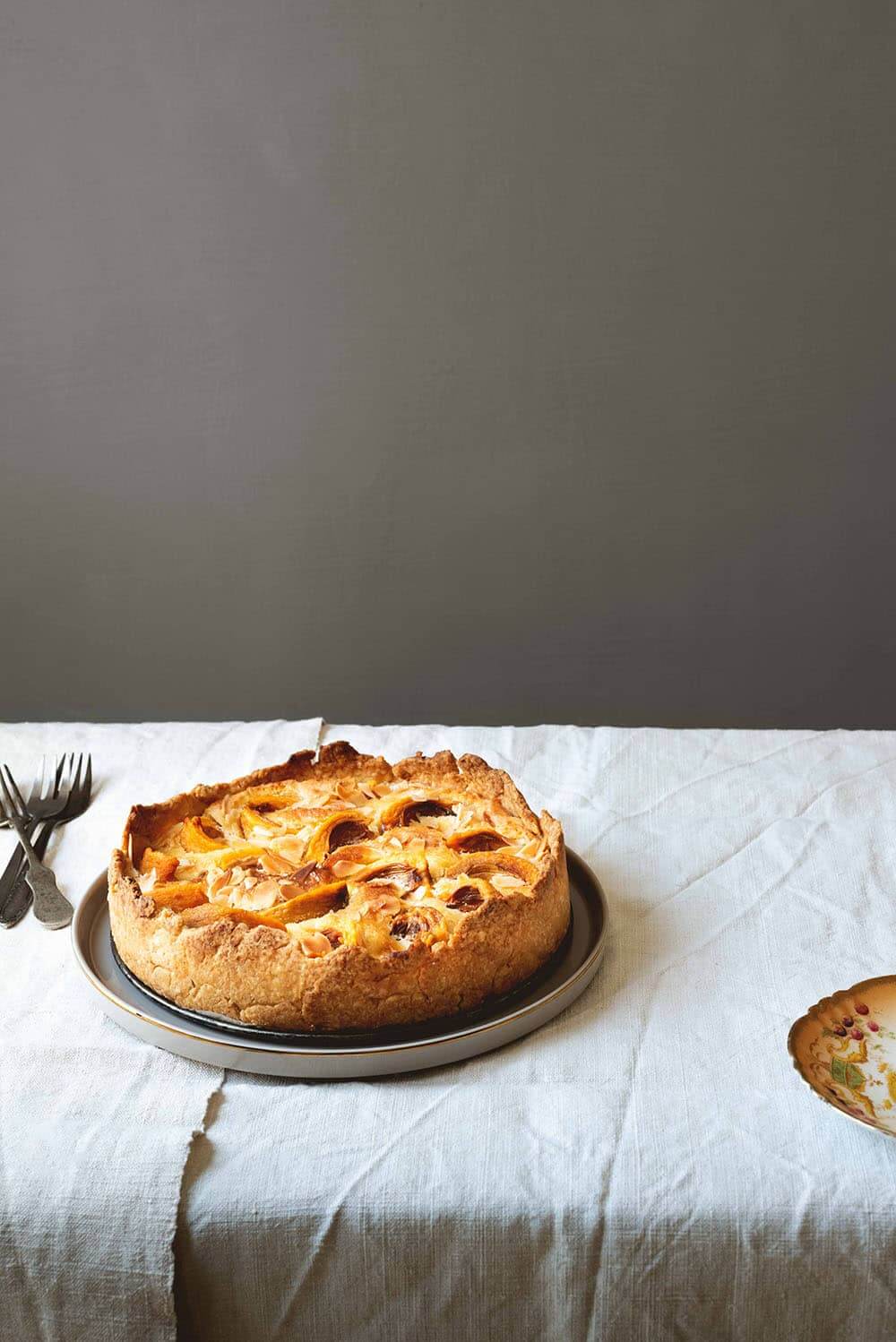 Tarta de nísperos En una mesa cubierta con un mantel blanco hay un plato gris. Encima está la tarta de nísperos. Alrededor hay cubiertos y un plato amarillo.