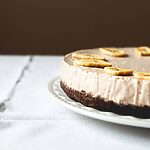 Una mesa gris está cubierta con un mantel blanco vintage. Encima está un plato blanco y encima está la tarta de plátano y queso. Está muy cerca.