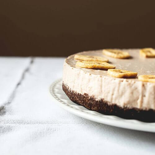 Una mesa gris está cubierta con un mantel blanco vintage. Encima está un plato blanco y encima está la tarta de plátano y queso. Está muy cerca.
