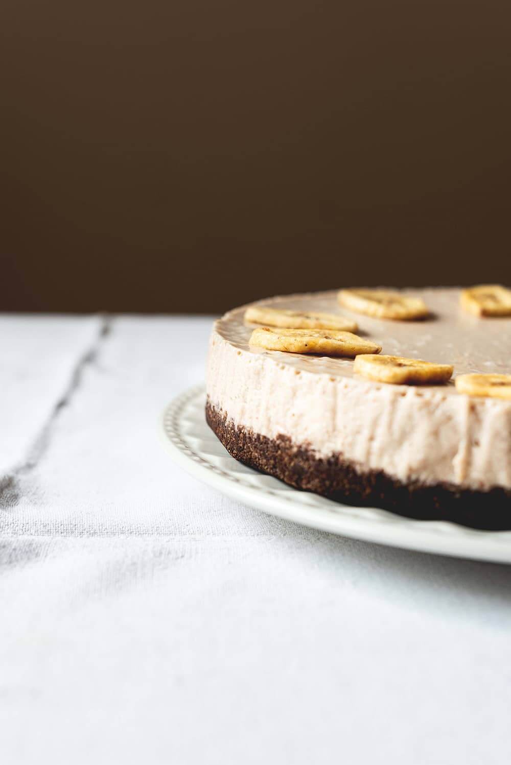Tarta de plátano y queso Una mesa gris está cubierta con un mantel blanco vintage. Encima está un plato blanco y encima está la tarta de plátano y queso. Está muy cerca.