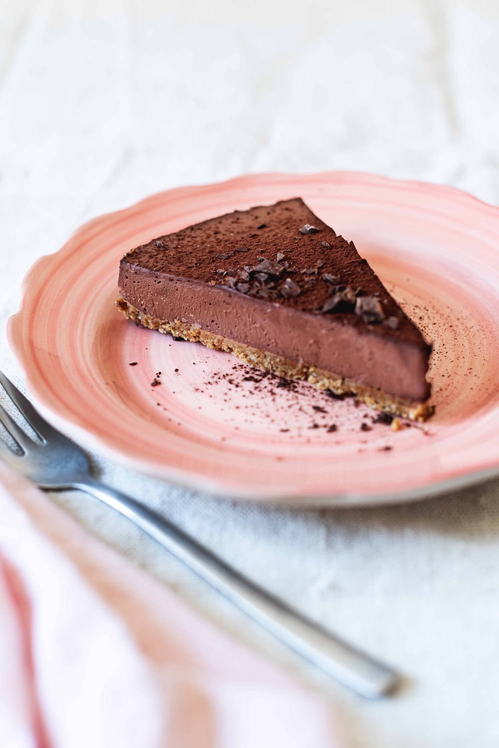 Tarta de queso de chocolate sin horno En una mesa gris está un plato rosa. Encima están dos raciones de la tarta de queso de chocolate sin horno. Al lado hay un tenedor y una servilleta rosa.