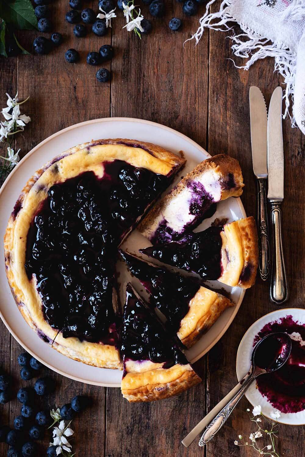 Tarta de queso y arándanos al horno En una mesa de madera hay un plato blanco. En el plato está la tarta de queso de arándanos. Está cortada y está cubierta con la sala de arándanos. Alrededor hay cubiertos de flores blancas, arándanos y hojas.
