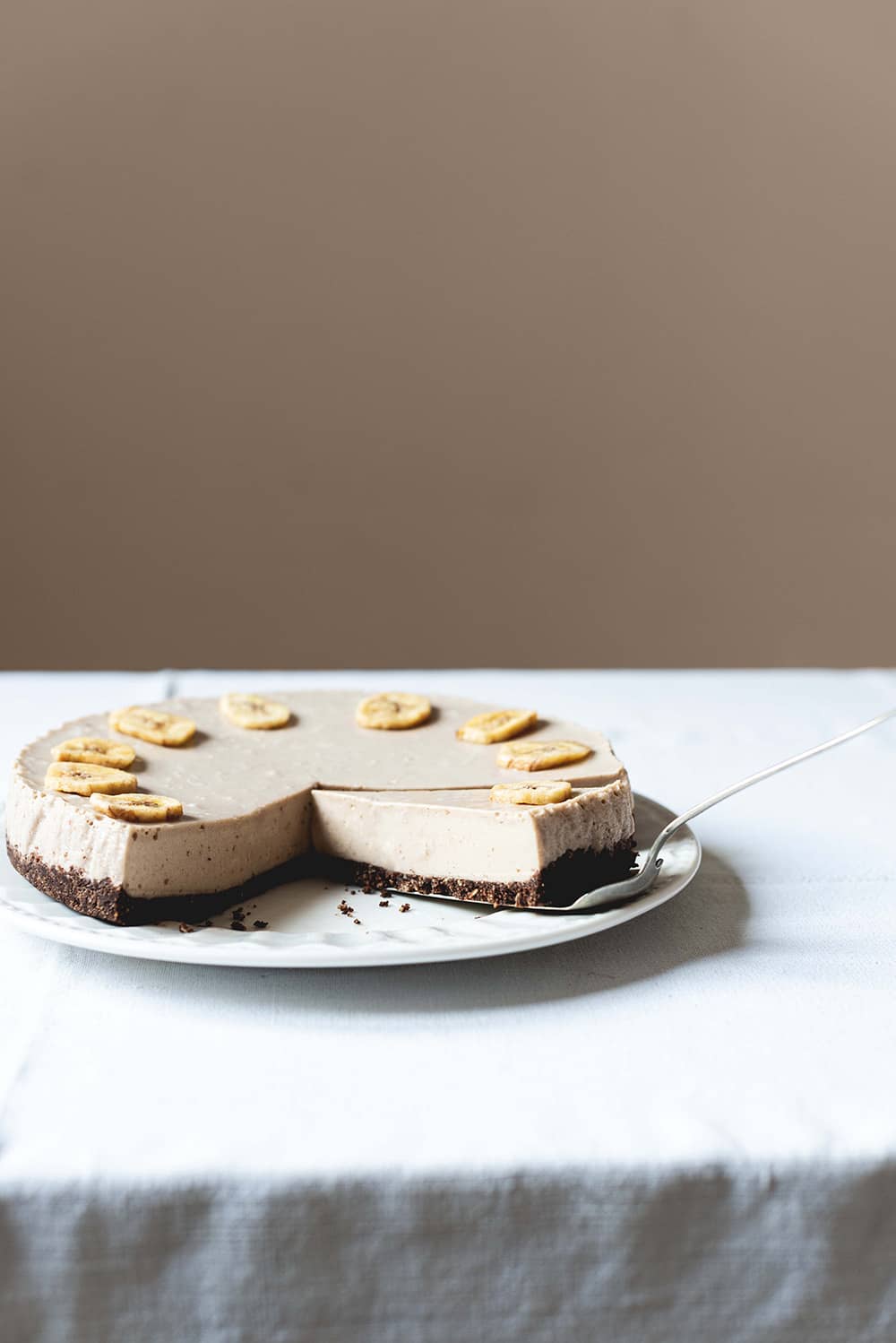 Tarta de queso y plátano sin horno Una mesa gris está cubierta con un mantel blanco vintage. Encima está un plato blanco y encima está la tarta de plátano y queso. Está cortada y se ve muy cremosa.