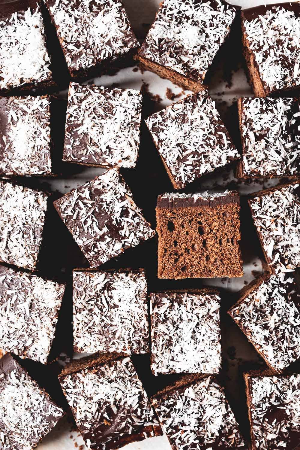 Encima de un papel de horno están los bizcochos de chocolate y coco ya cortados en cuadraditos. Se ven bien cerca, se nota que están esponjosos y se ve la capa a de crema chocolate y coco rallado encima de los cuadraditos.