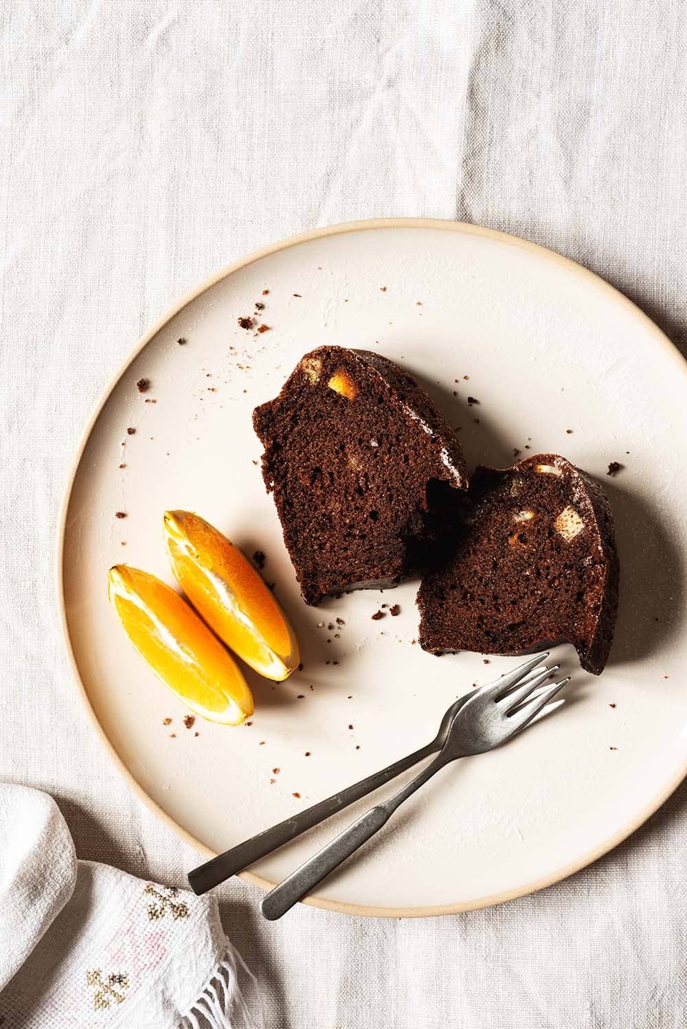 En una mesa está un mantel vintage. En el mantel hay un plato blanco. Encima hay dos raciones de bizcocho de chocolate con naranja. En el plato también hay dos gajos de naranjas y dos tenedores.