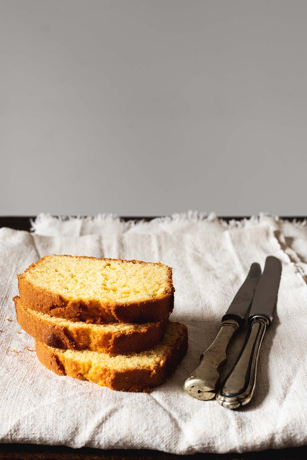 En una mesa de madera hay un mantel blanco. Encima del mantel están tres rodajas de Gateau weekend. Se ven muy tiernos y jugosos. Al lado del bizcocho hay dos cuchillos.