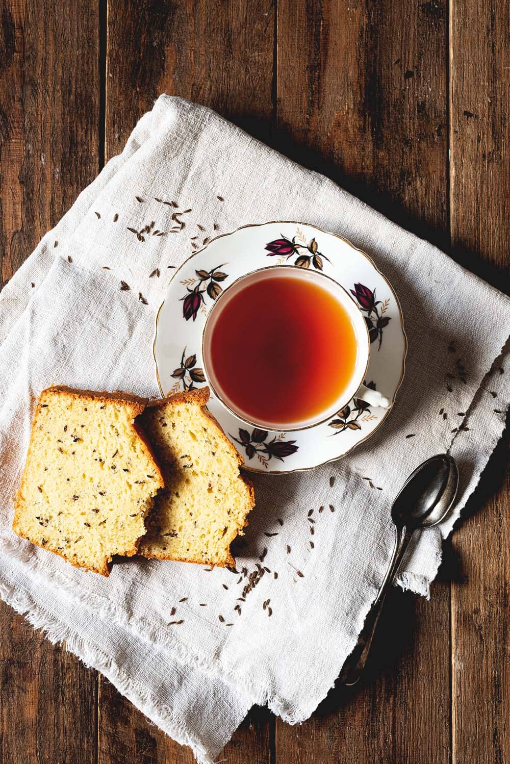 Se ve una superficie de madera. En un paño de cocina blanca están 2 rodajas de Bizcocho de semillas de alcaravea antiguo junto con una taza de té.