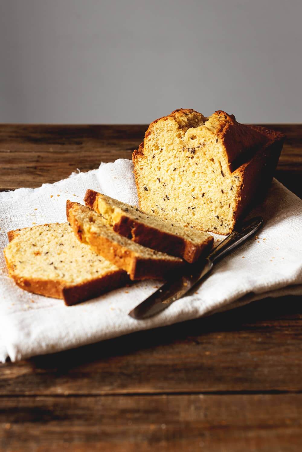 Se ve una superficie de madera. En un paño de cocina blanca está el Bizcocho de semillas de alcaravea esponjoso. Ya está cortado. Se ven migas suculentas y un cuchillo al lado de las rebanadas de bizcocho.