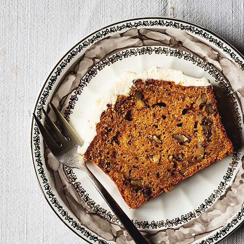 En una mesa hay un mantel blanco. Encima hay un plato vintage. En el plato hay una rodaja de bizcocho de zanahoria y nueces con su frosting de queso crema. Al lado del bizcocho hay un tenedor.