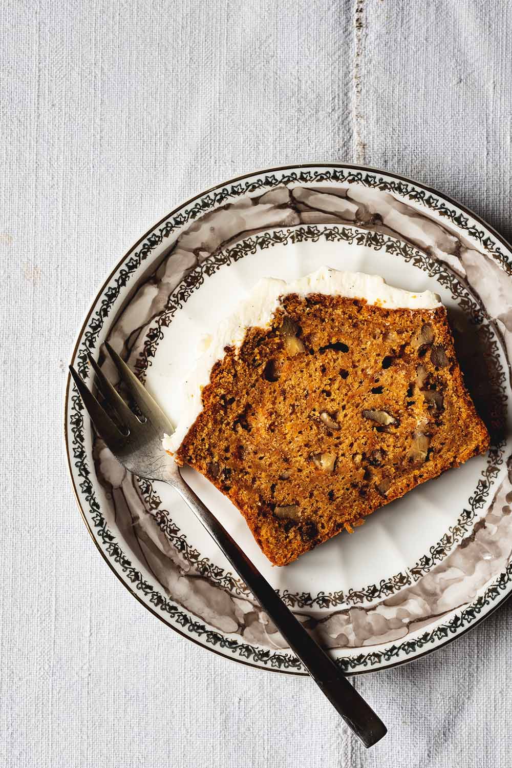 En una mesa hay un mantel blanco. Encima hay un plato vintage. En el plato hay una rodaja de bizcocho de zanahoria y nueces con su frosting de queso crema. Al lado del bizcocho hay un tenedor.