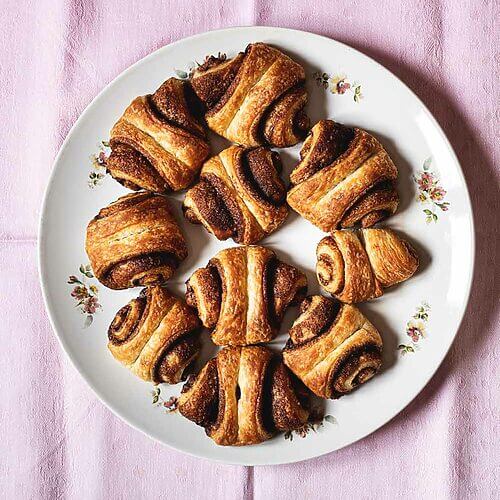 En un mantel de color rosa hay muchos Franzbrötchen. Se ven doraditos jugosos y llenos de relleno de canela.