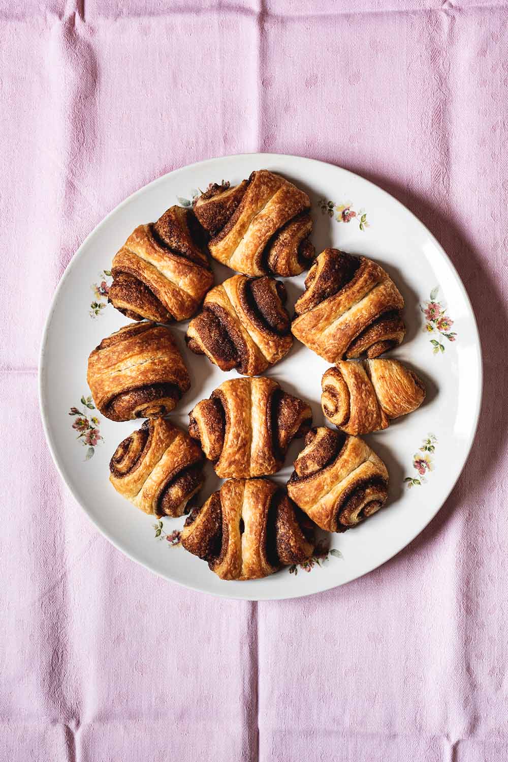En un mantel de color rosa hay muchos Franzbrötchen. Se ven doraditos jugosos y llenos de relleno de canela.