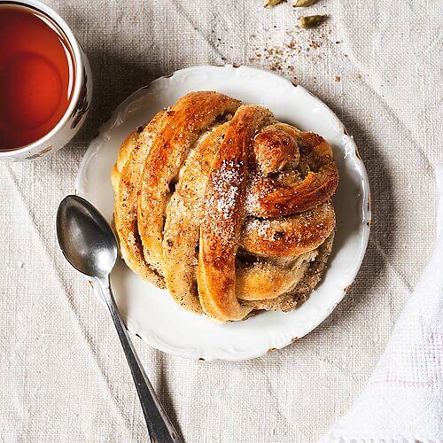 Bollos de cardamomo suecos esponjoso, recién horneado y espolvoreado con azúcar. Bollo trenzado junto a una taza de té caliente. Postre casero aromático y perfecto para acompañar el café o la merienda.