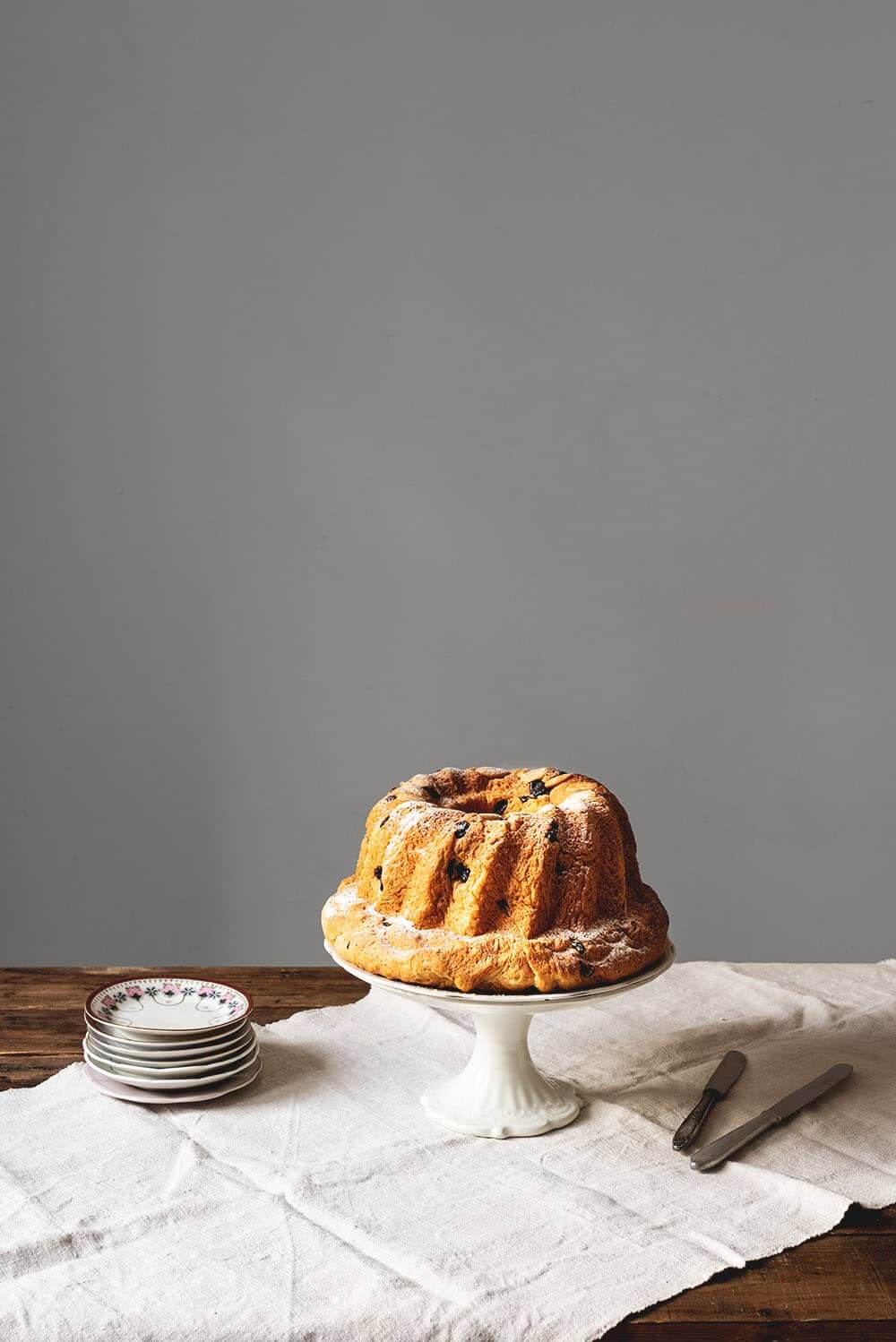 Brioche de Alsacia con pasas y almendras En una mesa de madera hay un mantel blanco. Encima del mantel hay un porta tartas. Encima está el Kouglof. Alrededor de porta tartas hay platos pequeños y cuchillos.