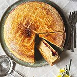 En el mantel hay un plato verde dónde está Pastel vasco tradicional ya cortado. Se ven las capas del postre. Crema pastelera, mermelada de cereza y masa quebrada. En el plato hay un colador pequeño con azúcar glas. También se ven al lado del postre un plato pequeño, flores y tenedores.