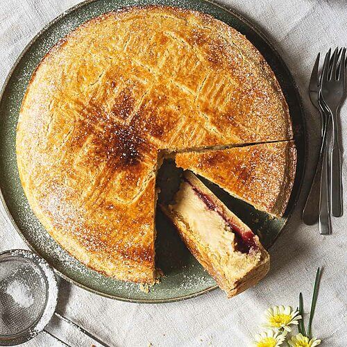 En el mantel hay un plato verde dónde está Pastel vasco tradicional ya cortado. Se ven las capas del postre. Crema pastelera, mermelada de cereza y masa quebrada. En el plato hay un colador pequeño con azúcar glas. También se ven al lado del postre un plato pequeño, flores y tenedores.