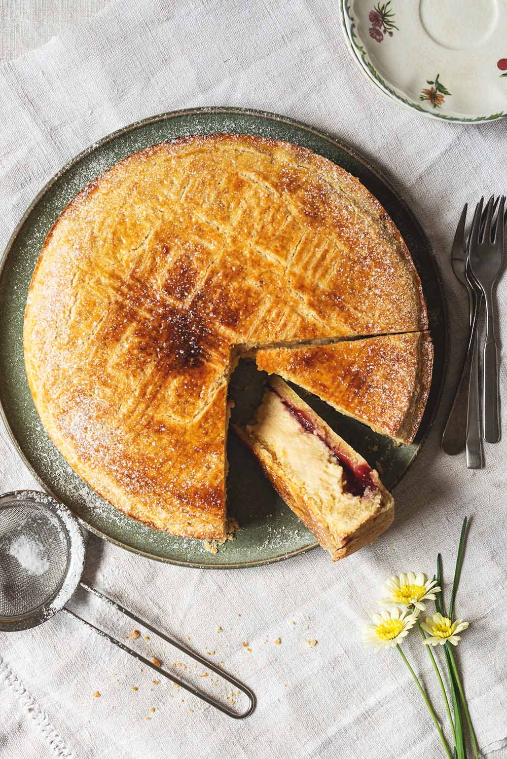 En el mantel hay un plato verde dónde está Pastel vasco tradicional ya cortado. Se ven las capas del postre. Crema pastelera, mermelada de cereza y masa quebrada. En el plato hay un colador pequeño con azúcar glas. También se ven al lado del postre un plato pequeño, flores y tenedores.