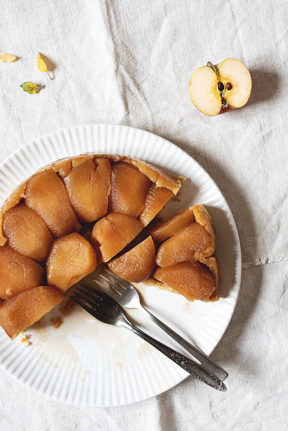 Se ve un mantel blanco tradicional. En el mantel está un plato grande balnco con una Tarta Tatin de manzana cortada. En plato hay dos tenedores. Alrededor del plato hay hojas y una manzana cortada en la mitad.